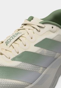 Κοντινό πλάνο του μπεζ και πράσινου αθλητικού παπουτσιού Adidas Adizero με κορδόνια, υφασμάτινο πάνω μέρος και τρεις χαρακτηριστικές ρίγες στο πλάι.