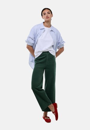 Pantalons en velours côtelé verts à jambes larges, t-shirt blanc et chemise oversize à rayures bleues et blanches. Ballerines rouges avec un accent en nœud.