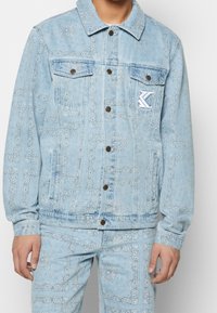 Light blue denim jacket with intricate white patterns, two chest pockets, button front, and a small white logo patch on the left side.