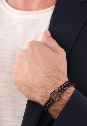 Brown braided leather bracelet featuring a square metallic accent with an engraved logo, worn on a wrist against a dark suit.
