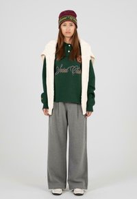 Groene polo met geborduurde tekst, gecombineerd met grijze wijde broek en een crèmekleurig gebreid vest. Bordeaurood hoed met logo-accenten.