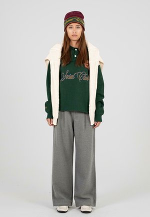 Groene polo met geborduurde tekst, gecombineerd met grijze wijde broek en een crèmekleurig gebreid vest. Bordeaurood hoed met logo-accenten.