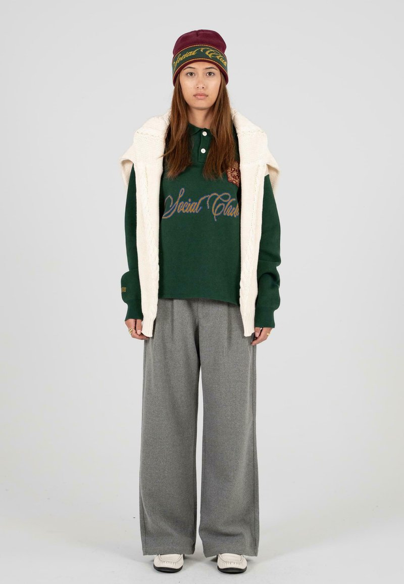 Groene polo met geborduurde tekst, gecombineerd met grijze wijde broek en een crèmekleurig gebreid vest. Bordeaurood hoed met logo-accenten.