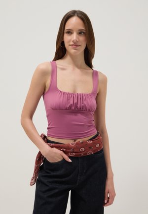 Junge Frau trägt ein ärmelloses mauvefarbenes gerafftes Croptop, schwarze Hosen und ein rotes gemustertes Halstuch, das um die Taille gebunden ist.