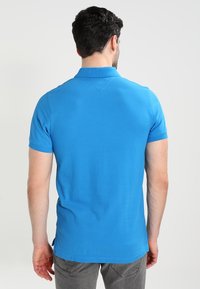 Polo bleu à manches courtes avec col, présentant une texture lisse et un ourlet droit. Porté avec un pantalon gris, vu de dos.