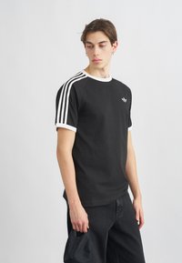 Čierne bavlnené tričko s bielymi akcentmi, s tromi bielymi pruhmi na rukávoch a malým logom na hrudi. Jednoduchý, okrúhly výstrih.