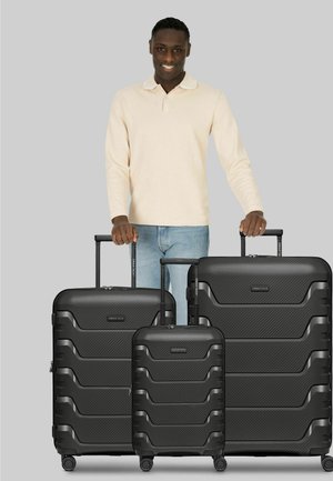 Uomo con camicia beige a maniche lunghe e jeans in piedi dietro tre valigie rigide nere con maniglie estese su sfondo grigio.