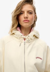 Haut à capuche zippé de couleur crème avec une doublure intérieure rose, cordon de serrage ajustable et logo rouge "SUPERDRY" sur la poitrine. Texture douce, coupe décontractée.