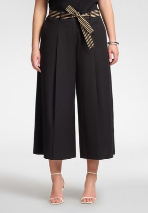 Frau trägt schwarze, weite, cropped Hosen mit einem gestreiften Gürtel, der zu einer Schleife gebunden ist, kombiniert mit weißen Riemchen-High Heels und einem schwarzen Oberteil.