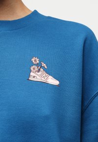 Μπλε φούτερ με ένα μικρό γραφικό ενός λευκού αθλητικού παπουτσιού που κρατά τρία λευκά λουλούδια μαργαρίτας στο στήθος.