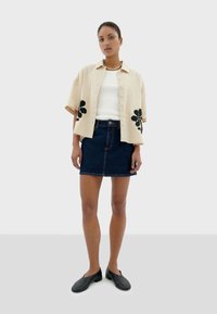 Beige short-sleeve button-up shirt with black floral accents, worn over a white top and paired with a dark denim mini skirt and black shoes.