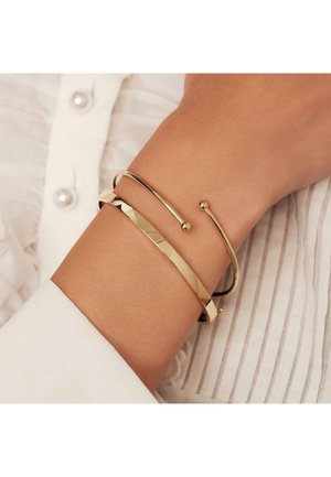 Des bracelets en or sur un poignet ; l'un lisse, l'autre texturé avec une légère courbure, présentant un accent en forme de boule. L'arrière-plan a une texture de tissu clair.