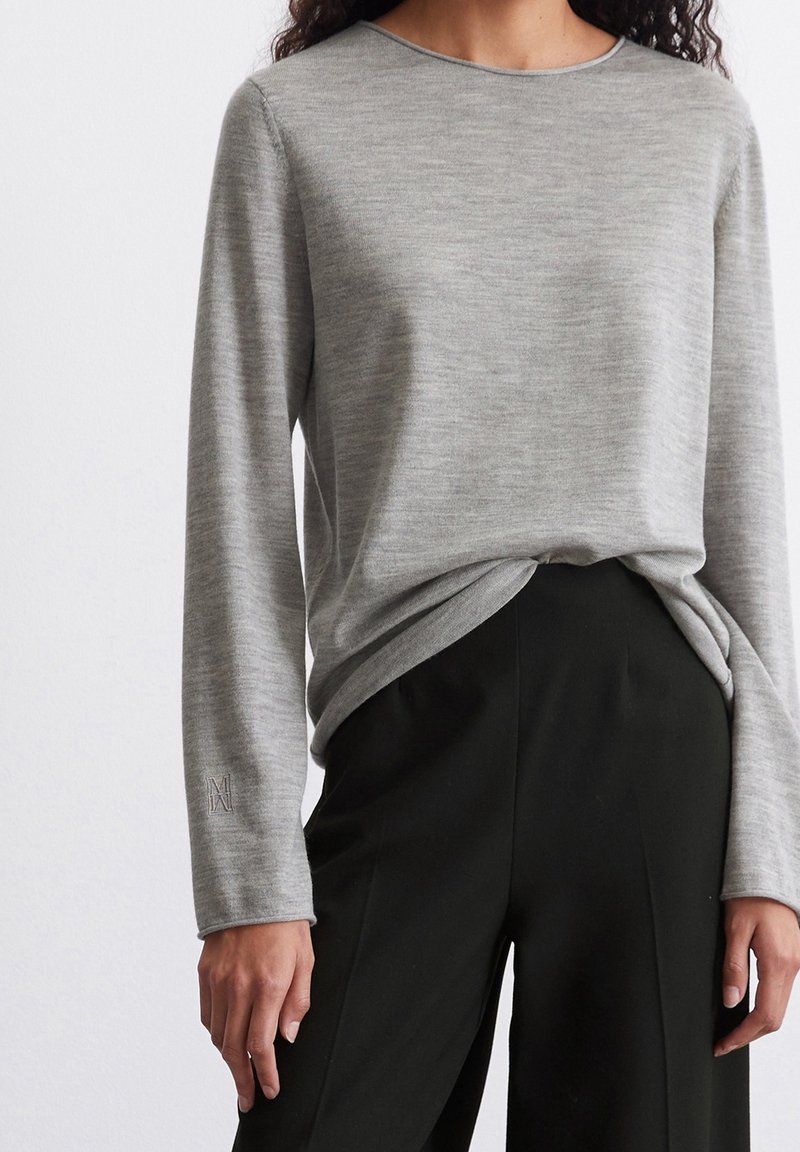 Pull gris clair à manches longues avec un col rond et un tissu texturé, présentant un détail de couture subtil au niveau du poignet, assorti à un pantalon noir.