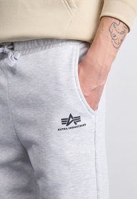 Lys grå joggebukser laget av mykt stoff med en sidelomme og en svart "Alpha Industries"-logo på låret.