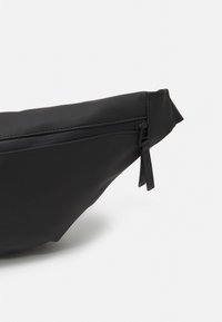 Černá ledvinka vyrobená z měkkého materiálu, s zipovým uzávěrem, hladkým povrchem a minimalistickým, zakřiveným designem.