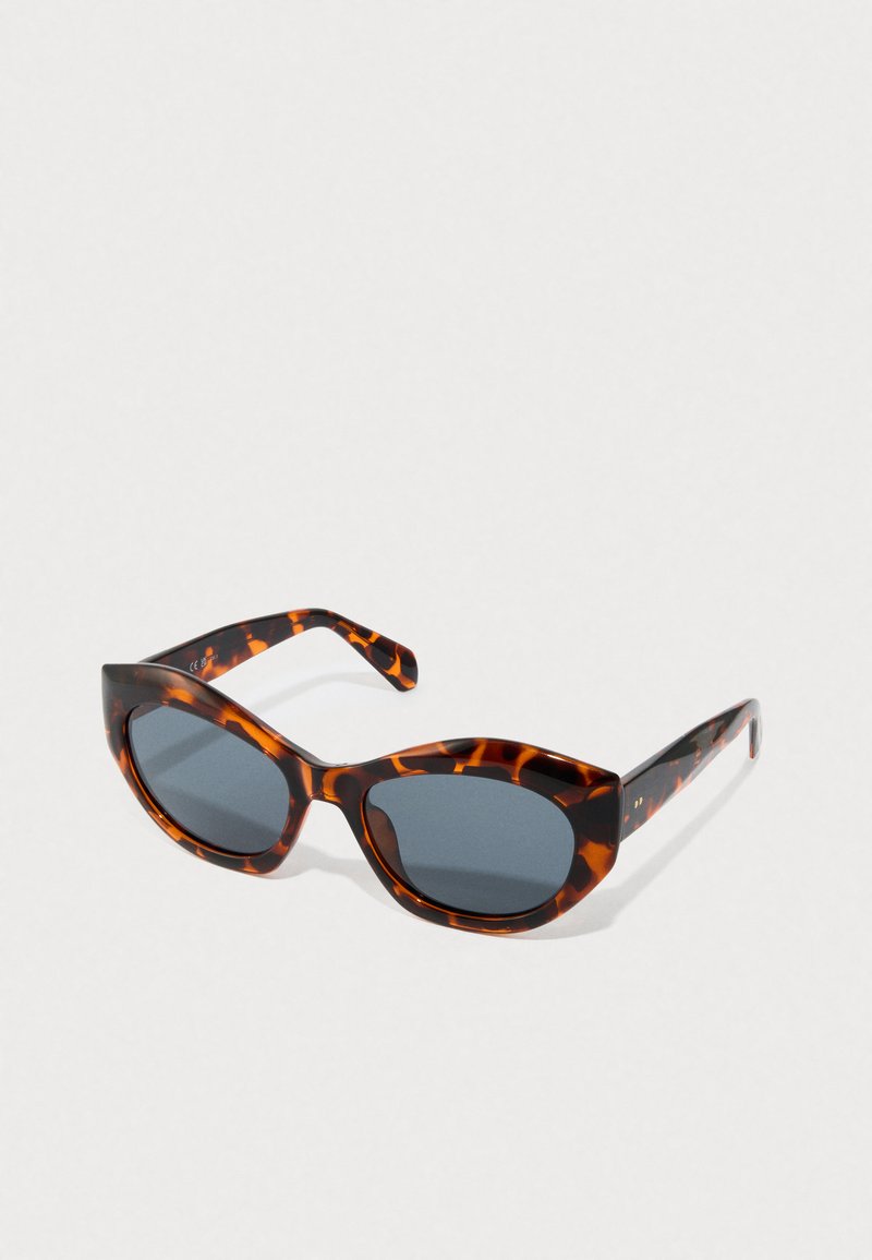 Occhiali da sole a forma di occhio di gatto in tartaruga con lenti scure, poggiati su una superficie chiara e semplice.