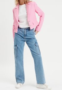 Blazer rose texturé, t-shirt blanc, jean bleu à jambes larges avec poches cargo et baskets blanches. Le blazer est doté de boutons argentés et de revers à encoche.