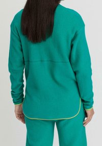 Maglione in pile turchese con maniche lunghe, caratterizzato da un orlo curvo e accenti gialli. Tessuto morbido e vestibilità rilassata.