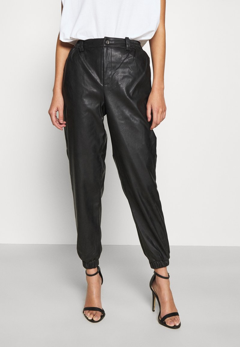 Kvinna som bär högmidjade svarta faux läder joggerbyxor med elastiska muddar, kombinerat med svarta högklackade sandaler med ankelrem.