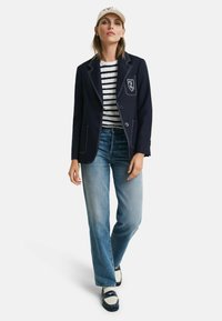 Blazer marine avec des coutures blanches et un insigne, chemise rayée, jeans bleu clair et mocassins bleu marine et blancs, complétés par une casquette beige.