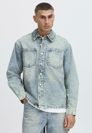 Giovane uomo con giacca di jeans blu chiaro effetto acid wash e jeans abbinati, con camicia bianca sotto, in piedi davanti a uno sfondo neutro.