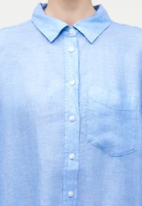 Prim-plan al unei cămăși bleu deschis cu nasturi, cu guler și buzunar la piept, purtată de o persoană.