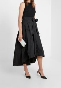 Czarna bezrękawnik z teksturowanym wykończeniem, zestawiony z zwiewną czarną spódnicą z wiązaniem w talii. Uzupełniony srebrną kopertówką.