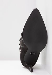 Chaussure à talon haut en daim noir à bout pointu vue de la semelle, avec un talon stiletto fin et des accents de boucle argentée.