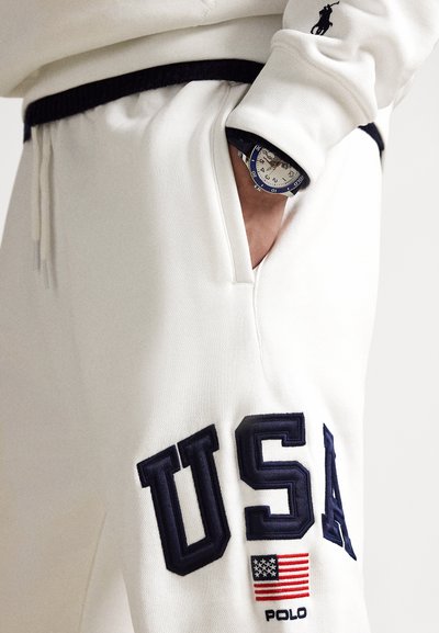 Biele teplákové nohavice z textúrovanej, mäkkej látky s veľkou námorníckomodrou výšivkou "USA" a detailom americkej vlajky. Súčasťou je tmavý pás a vrecko.
