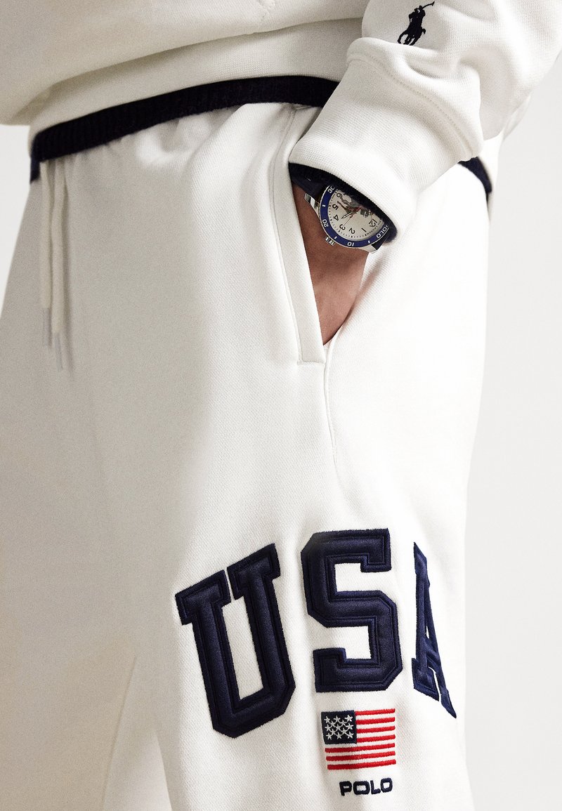 Pantalons de jogging blancs en tissu doux et texturé, arborant une grande broderie "USA" en bleu marine et un détail du drapeau américain. Comprend une taille foncée et une poche.