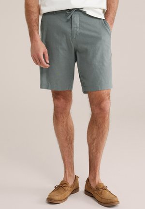 Homme portant un short gris à cordon et des mocassins en daim beige, debout avec une main dans la poche sur fond neutre.