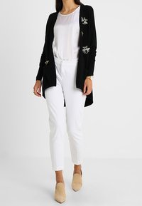 Svart cardigan med broderade bi-motiv, bärs över en vit blus och vita byxor. Beiga spetsiga skor fulländar outfiten.