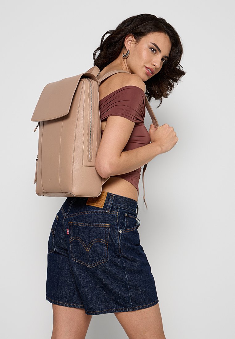 Beige Leder-Rucksack mit einem Klappenverschluss und vertikalem Reißverschluss. Glatte Textur, rechteckige Form, einseitig auf der Schulter getragen. Denim-Shorts sichtbar.