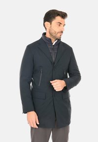 Cappotto ibrido blu scuro con rever a intaglio, tasche frontali e tasca sul petto con cerniera, realizzato in materiale liscio e resistente all'acqua.