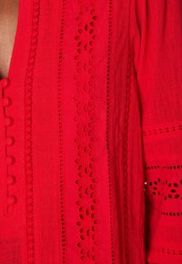 Blusa rossa ricamata con dettagli decorativi in bottoni e intagli in pizzo. Tessuto testurizzato e ricami intricati lungo la parte anteriore e le maniche.