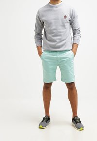 Mann in einem grauen Langarmshirt, hellblauen Shorts und grauen Turnschuhen mit neon-grünen Akzenten, der mit den Händen in den Taschen dasteht.
