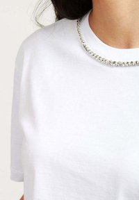 T-shirt in cotone bianco con scollo rotondo adornato da una fila di piccoli strass trasparenti, che creano un sottile accento decorativo.