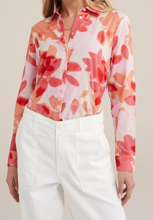 Vrouw draagt een blouse met lange mouwen en knopen met een rood en oranje bloemenpatroon, ingestopt in een witte broek met hoge taille en voorzakken.