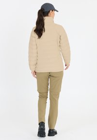 Beige vadderad jacka med hög krage, kombinerad med khakifärgade byxor. Modellen bär en mörk keps och svarta vandringskängor. Enkel och funktionell design.