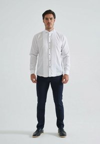 Chemise blanche avec un devant plissé, col haut, fermeture à boutons et boutons noirs. Associée à un pantalon bleu foncé et des chaussures noires.