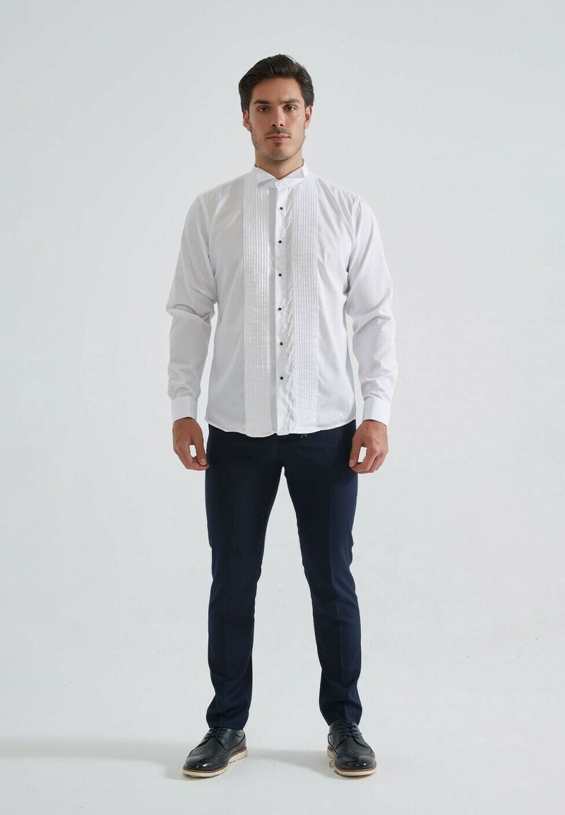 Chemise blanche avec un devant plissé, col haut, fermeture à boutons et boutons noirs. Associée à un pantalon bleu foncé et des chaussures noires.
