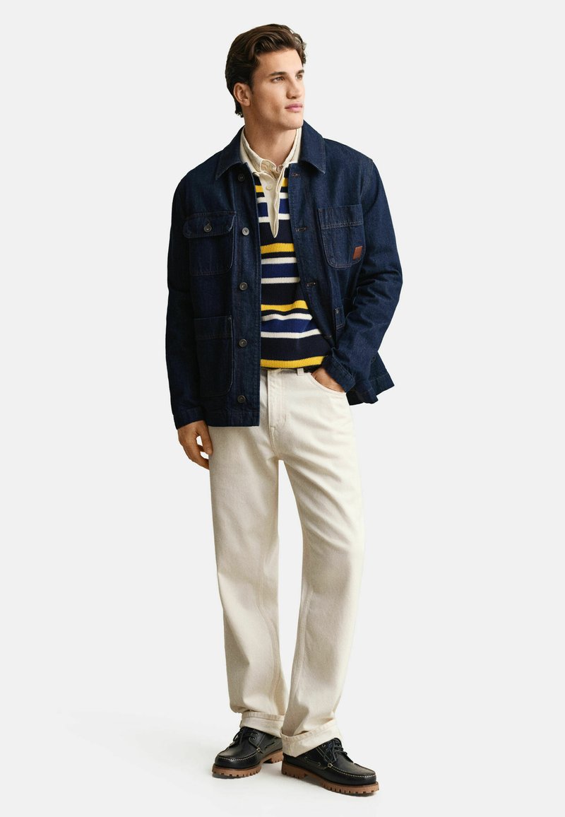 Jeune homme portant une veste bleu marine, un pull rayé, un pantalon beige et des chaussures noires, debout avec une main dans la poche, regardant vers la droite.