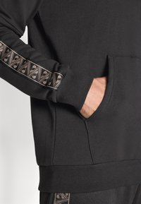 Zwart sweatshirt van structuurtstof, met een geometrisch patroon in koper langs de mouw. Inclusief een voorvak en ribgebreide boorden.