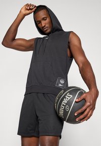 Sudadera sin mangas negra con un bolsillo frontal y detalle de logo, combinada con pantalones cortos de malla negros. El modelo sostiene un balón de baloncesto negro.