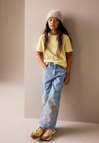 Gul t-shirt med korte ærmer lag på en brun langærmet skjorte, blå jeans med farverige smiley-designs og flerfarvede sneakers.