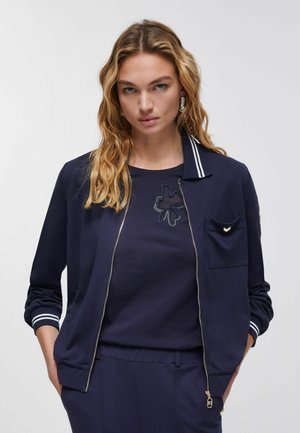 Donna con lunghi capelli biondi che indossa una giacca blu marino con cerniera sopra una maglietta e pantaloni abbinati, in piedi con le mani nelle tasche.