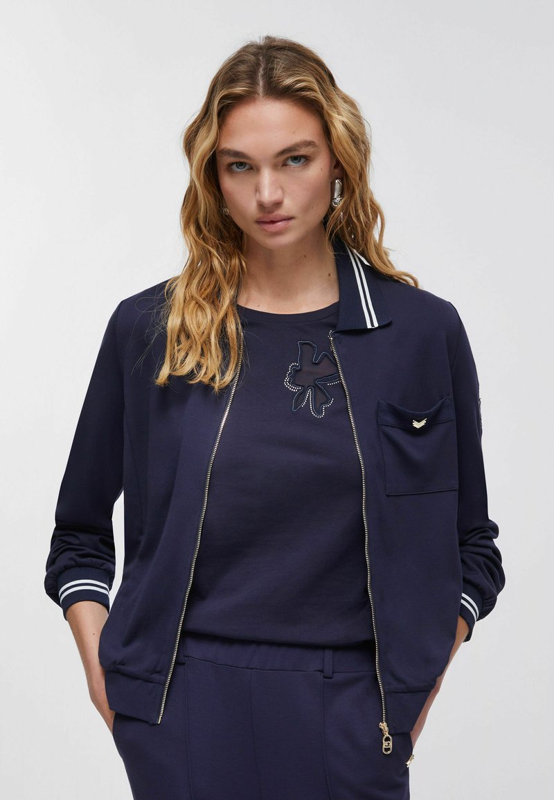 Femme aux longs cheveux blonds portant une veste zippée bleu marine sur un ensemble chemise et pantalon assorti, debout les mains dans les poches.