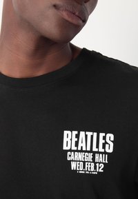 Black cotton t-shirt featuring white printed text that reads "BEATLES CARNEGIE HALL WED. FEB.12" with additional concert details below.