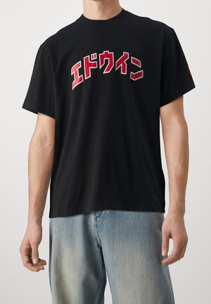 Mężczyzna w czarnym T-shircie z krótkim rękawem z wyraźnym czerwono-białym japońskim napisem na piersi oraz jasnoniebieskich dżinsach.