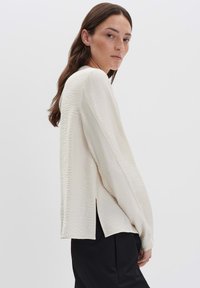 Donna in una blouse crema a maniche lunghe con texture e aperture laterali, abbinata a pantaloni neri a vita alta, in piedi su uno sfondo semplice.